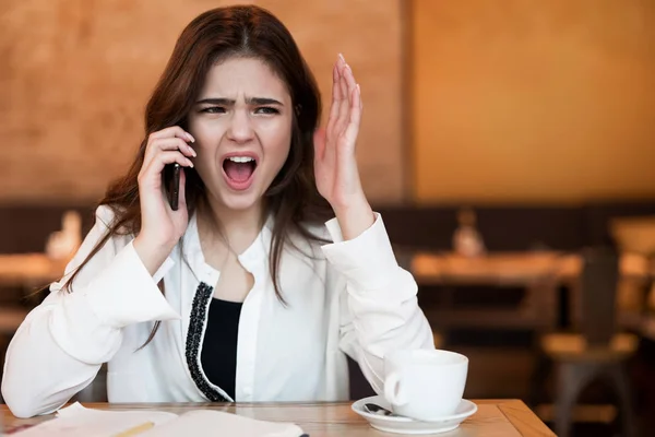Mujer joven en los teléfonos aéreos que tienen una conversación telefónica desagradable trabajando fuera de la oficina en su computadora portátil durante la pausa para comer beber café caliente en la cafetería mirando irritado — Foto de Stock