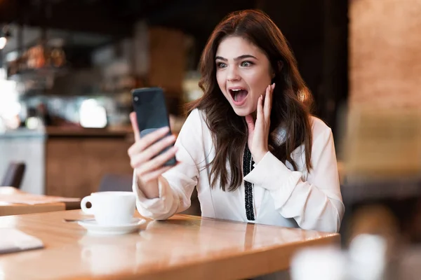 Joven hermosa mujer en chaqueta blanca con estilo con teléfono inteligente en sus manos mirando sorprendido mientras habla con un amigo durante el descanso de café en la cafetería moderna mujer de negocios — Foto de Stock