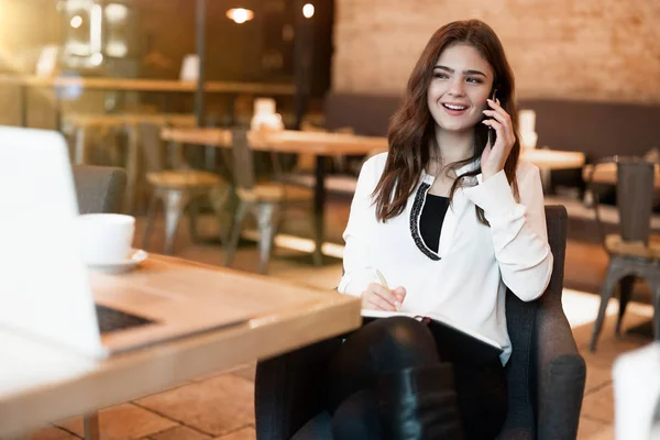 Joven hermosa mujer en chaqueta blanca con estilo con teléfono inteligente en sus manos mirando complacido mientras habla con un amigo durante el descanso de café en la cafetería moderna mujer de negocios — Foto de Stock