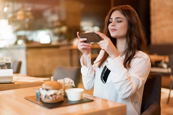 Joven hermosa mujer en blusa blanca con estilo tomar una foto de su comida hamburguesa de carne mientras come el almuerzo en la cafetería de moda moderna mujer de negocios multitarea — Foto de Stock