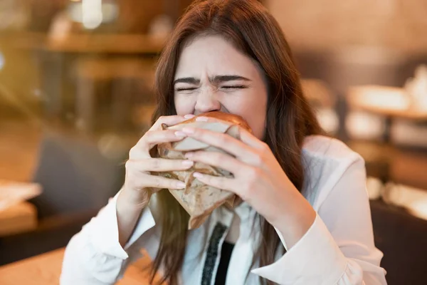 Mujer joven en blusa blanca y elegante mordiendo con apetito hamburguesa de carne fresca durante el almuerzo en un restaurante de moda mirando hambriento comiendo al aire libre —  Fotos de Stock
