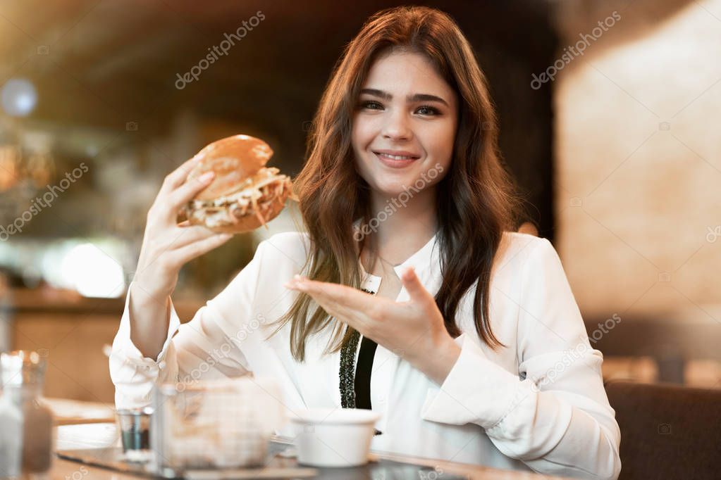 young beautiful woman in white stylish blouse looks hungry pointing with her hand at fresh meat burger during lunch in trendy cafe eating outside
