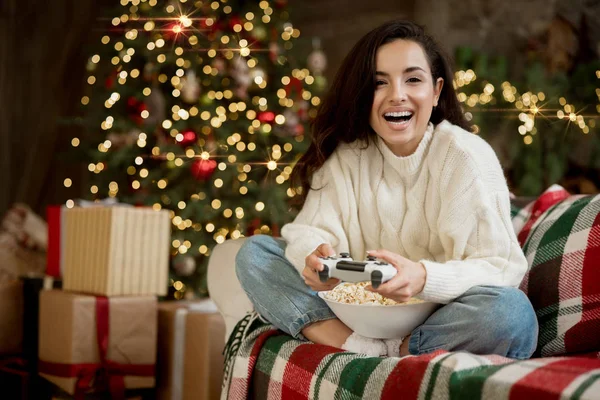 Morena mujer vistiendo suéter jugando videojuego en el sofá con palomitas de maíz en la habitación decorada para celebrar el año nuevo y el estado de ánimo festivo de Navidad —  Fotos de Stock