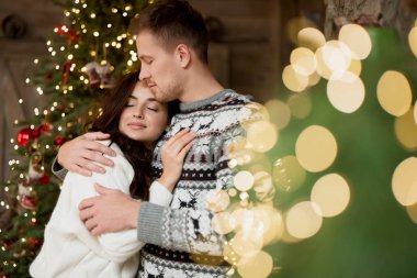Güzel bir çift, güzel karısına sarılıyor. Noel ağacının yanında duruyor. Yeni yılı kutlamak için dekore edilmiş bir odada mutlu görünüyor.