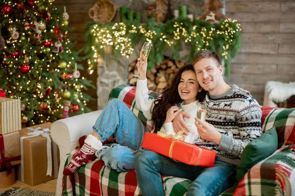 lovely couple man and woman both wearing warm sweaters hugging on sofa in room decorated for celebrating the new year christmas drinking champagne love story