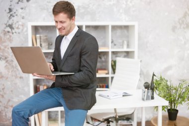 Dizüstü bilgisayarda çalışan gülümseyen yakışıklı yönetici ofisteki iş yerinde masanın üzerinde mutlu görünüyor. Çoklu görev, çalışma konsepti