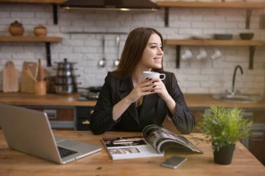 smiling beautiful business woman checking daily news in laptop and reading magazine in the kitchen while drinking morning coffee before going to office . multitasking, rituals clipart