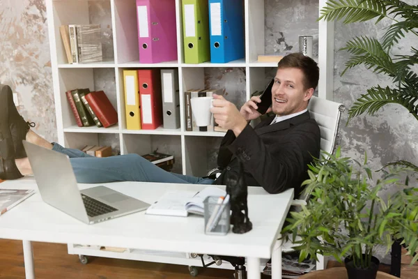 Handsome man manager sitting at his working place with his legs on the table drinking coffee having phone conversation looking happy — Stock Photo, Image