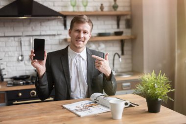 Genç işadamı, ofise gitmeden önce mutfakta dergi okurken modern akıllı telefonunu işaret ederek sabah kahvesini içiyor. Çoklu görev, sabah