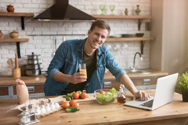 Молодой улыбающийся человек со стаканом свежего сока приготовление завтрака на кухне проверка рецепт в своем ноутбуке, в воскресенье утром — стоковое фото