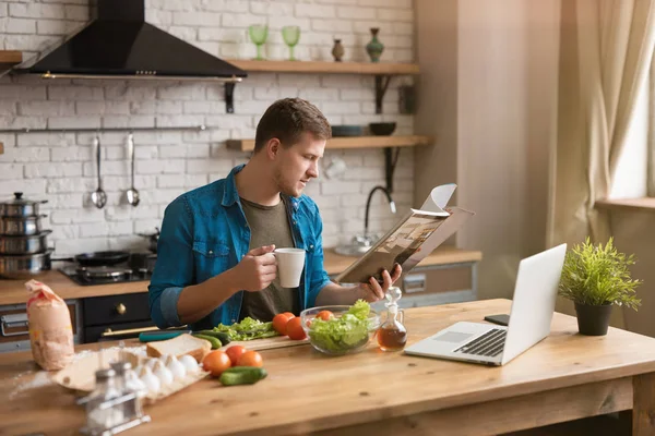 Ung leende man läser tidningen medan du äter frukost i välutrustat kök kontroll recept i sin bärbara dator, söndag morgon — Stockfoto