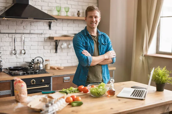 Ung leende man står i välutrustat kök samtidigt som frukost kontroll recept i sin bärbara dator, söndag morgon — Stockfoto