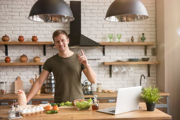 Ung leende man stående i välutrustat kök letar efter intressanta idéer för frukost kontroll recept i sin bärbara dator, söndag morgon — Stockfoto