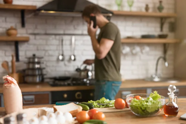 Produkter till frukost på bordet: tomat, ägg, olivolja, gurka, sallad man står nära spis talar i telefonen på suddig bakgrund — Stockfoto