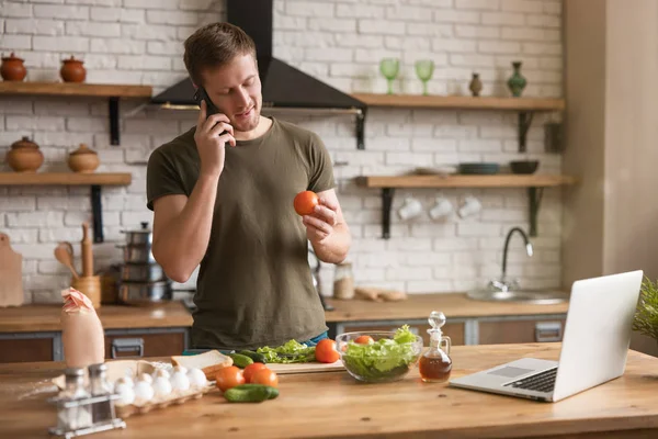 Ung leende man talar i telefon med tomat i sin habd medan matlagning frukost i köket kontroll recept i sin bärbara dator, söndag morgon — Stockfoto