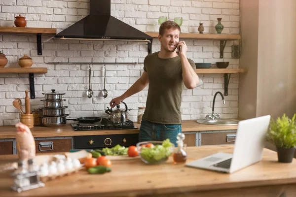 Ung leende man står nära spis i välutrustat kök med telefon samtal letar efter intressanta idéer för frukost kontroll recept i sin bärbara dator, söndag morgon — Stockfoto