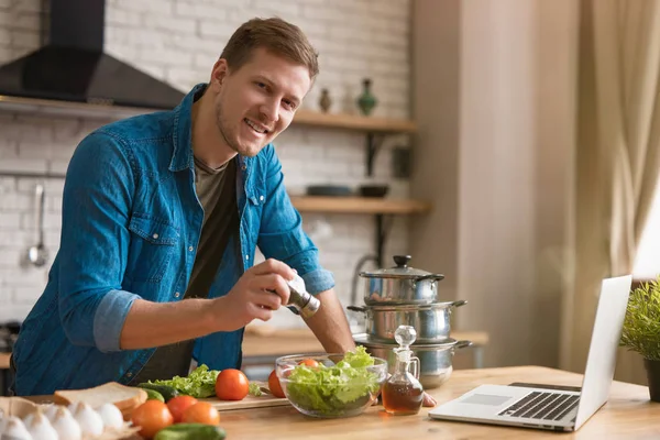 Ung stilig man lägga kryddor till grönsaker samtidigt förbereda sallad för lunch stående i välutrustat modernt kök kontroll recept i laptop, helg vibbar — Stockfoto