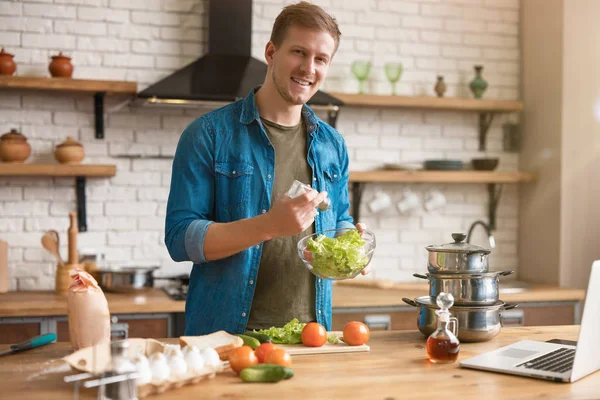 Ung stilig man lägga till salt och kryddor till sallad medan matlagning lunch stående i välutrustat modernt kök kontroll recept i laptop, helg vibbar — Stockfoto