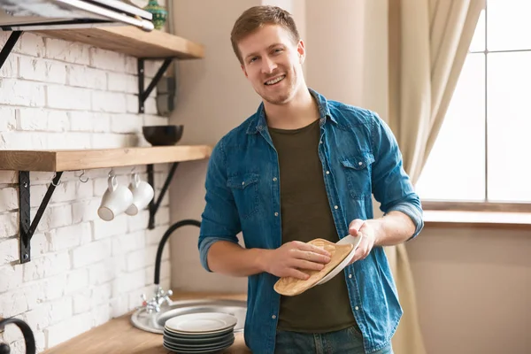 Joven hombre sonriente de pie en brillante cocina moderna bien equipada limpiando platos limpios con toalla después de lavar, el domingo por la mañana vibraciones — Foto de Stock
