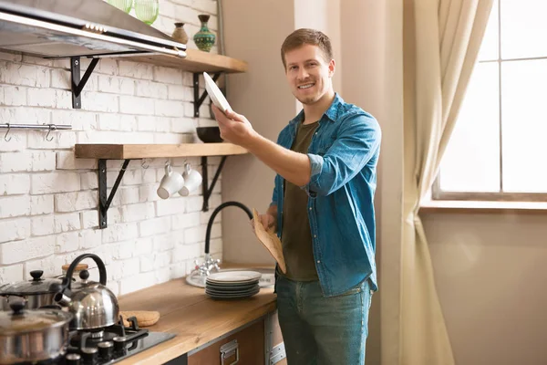 Joven hombre sonriente de pie en brillante cocina moderna bien equipada limpiando platos limpios con toalla después de lavarlos y ponerlos en el estante, la vida de soltero — Foto de Stock