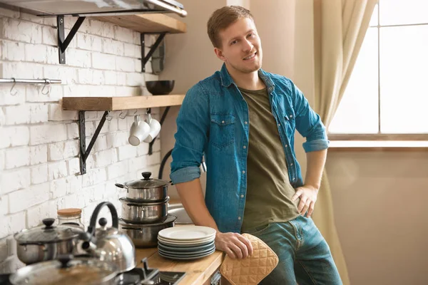 Ung leende man står i ljusa välutrustade modernt kök ser nöjd efter diska, ungkarlsliv — Stockfoto