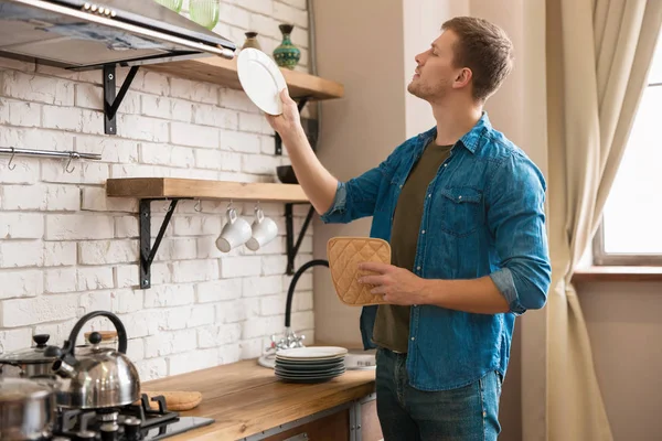 Joven hombre guapo de pie en brillante cocina moderna bien equipada sosteniendo plato limpio mientras se limpian los platos con toalla después de lavar, el domingo por la mañana vibraciones — Foto de Stock