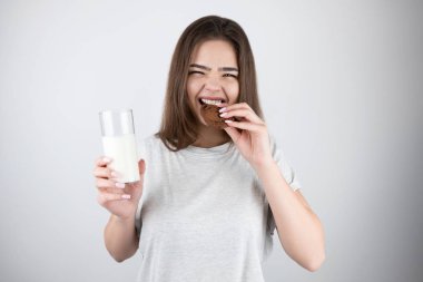 Genç, güzel esmer kadın çikolatalı kurabiyeyi ısırıyor. Elinde bir bardak süt tutuyor. Sağlıklı yaşam tarzı izole edilmiş beyaz arka plan.