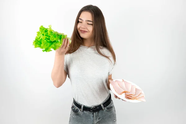 Ung vacker leende brunett kvinna håller färsk sallad i ena handen och platta med skinka i en annan ser glad hälsosam livsstil isolerad vit bakgrund — Stockfoto