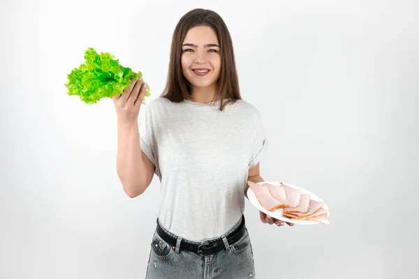 Vacker passform brunett kvinna håller färsk sallad i ena handen och platta med skinka i en annan ser glad ut att vara på diet isolerad vit bakgrund — Stockfoto