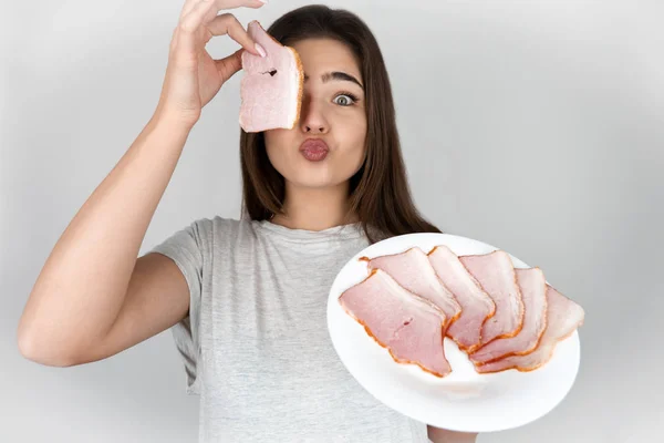 Wanita cantik muda memegang sepotong ham di satu tangan dan piring dengan ham di lain tampak lucu berada di diet terisolasi latar belakang putih — Stok Foto