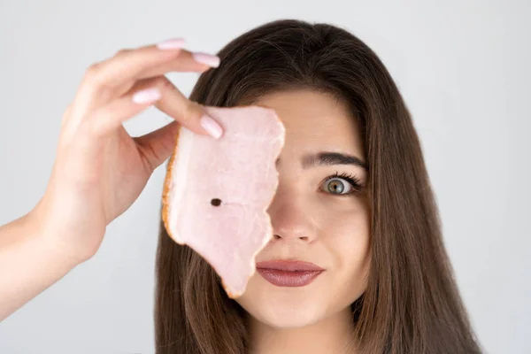 Jong mooi grappig vrouw holding slice van ham in de buurt van haar oog op zoek hongerig geïsoleerd wit achtergrond — Stockfoto