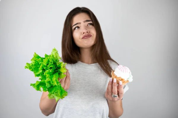 Jovem bela mulher segurando salada verde em uma mão e cupcake doce com creme em outra olhando frustrado estar na dieta isolado fundo branco — Fotografia de Stock