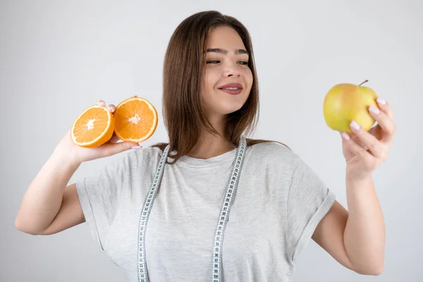 Jovem mulher com centímetro em torno de seu pescoço olhando para maçã em sua mão wih duas metade de laranjas em outro fundo branco isolado dietologia nutrição — Fotografia de Stock