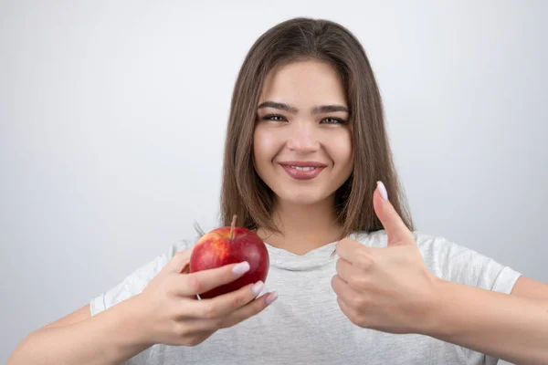 Jovem morena sedutora mulher segurando maçã vermelha na mão mostrando como sinal de pé sobre fundo branco isolado dietologia e nutrição — Fotografia de Stock