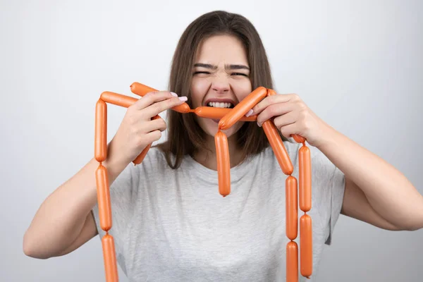 Jong mooi glimlachende vrouw houden vlees worsten in beide handen bijten ze met honger staan op geïsoleerde witte achtergrond dieet en voeding — Stockfoto