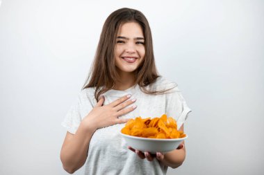 Elinde baharatlı patates cipsi olan genç, çekici, mutlu bir kadın izole edilmiş beyaz arka plan diyetolojisi ve beslenme şekline şaşırmış görünüyor.