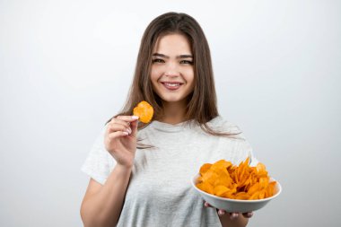Genç ve çekici bir kadın elinde bir parça cips ile izole edilmiş beyaz arka plan dietolojisi ve beslenme üzerine baharatlı patates cipsi tabağıyla ayakta duruyor.