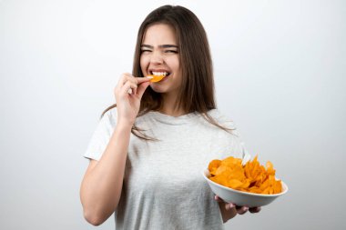 Genç ve çekici bir kadın açlıkla ısırıyor elinde bir parça cips izole edilmiş beyaz arka plan dietolojisinde baharatlı patates cipsi tabağıyla duruyor.