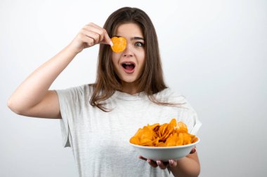 Genç ve çekici bir kadın, gözünün yanında bir parça cips tutuyor. İzole edilmiş beyaz arka plan dietolojisi ve beslenme üzerine baharatlı patates cipsi tabağıyla ayakta duruyor.