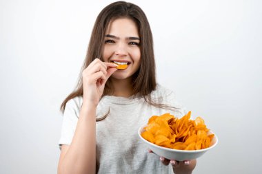Genç ve çekici bir kadın ağzına bir parça cips koyuyor. İzole edilmiş beyaz arka plan dietolojisi ve beslenme üzerine baharatlı patates cipsi ile birlikte duruyor.
