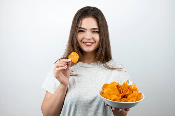 Jonge aantrekkelijke vrouw met een stuk chips in haar hand staan met plaat van pittige aardappel chips op geïsoleerde witte achtergrond dieet en voeding — Stockfoto