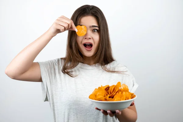 Ung attraktiv kvinna som håller en bit chips nära ögat stående med tallrik kryddig potatischips på isolerad vit bakgrund dietologi och näring — Stockfoto