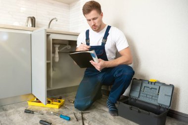 Genç yakışıklı tesisatçı, mutfak tamirhanesinde boruları tamir ederken talimatları kontrol ederken konsantre olmuş görünüyor.