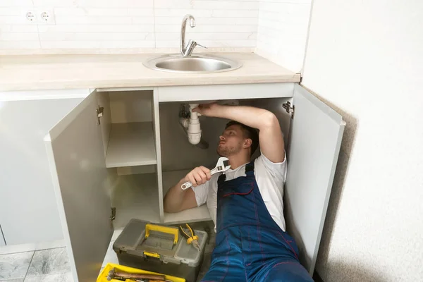 Fontanero joven en uniforme fijación del fregadero con llave ajustable en su mano acostado en el piso de la cocina servicio de reparación de fontanería profesional — Foto de Stock