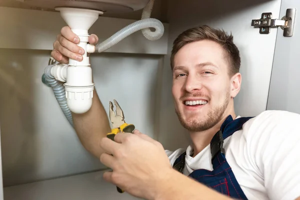 Joven guapo sonriente fontanero en uniforme fijar el fregadero con alicates en la mano sentado en el piso de la cocina servicio de reparación de fontanería profesional — Foto de Stock