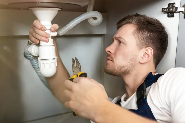 Joven hombre guapo fontanero en uniforme fijación del fregadero con alicates en la mano sentado en el piso de la cocina servicio de reparación de fontanería profesional — Foto de Stock