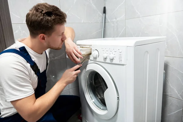 Young handsome man worker in uniform repairing washing machine at home in the toilette professional repair service — Stock Photo, Image