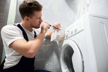 Evde tuvalette çamaşır makinesini tamir eden üniformalı genç yakışıklı adam yoğun profesyonel tamir servisi gibi görünüyor.