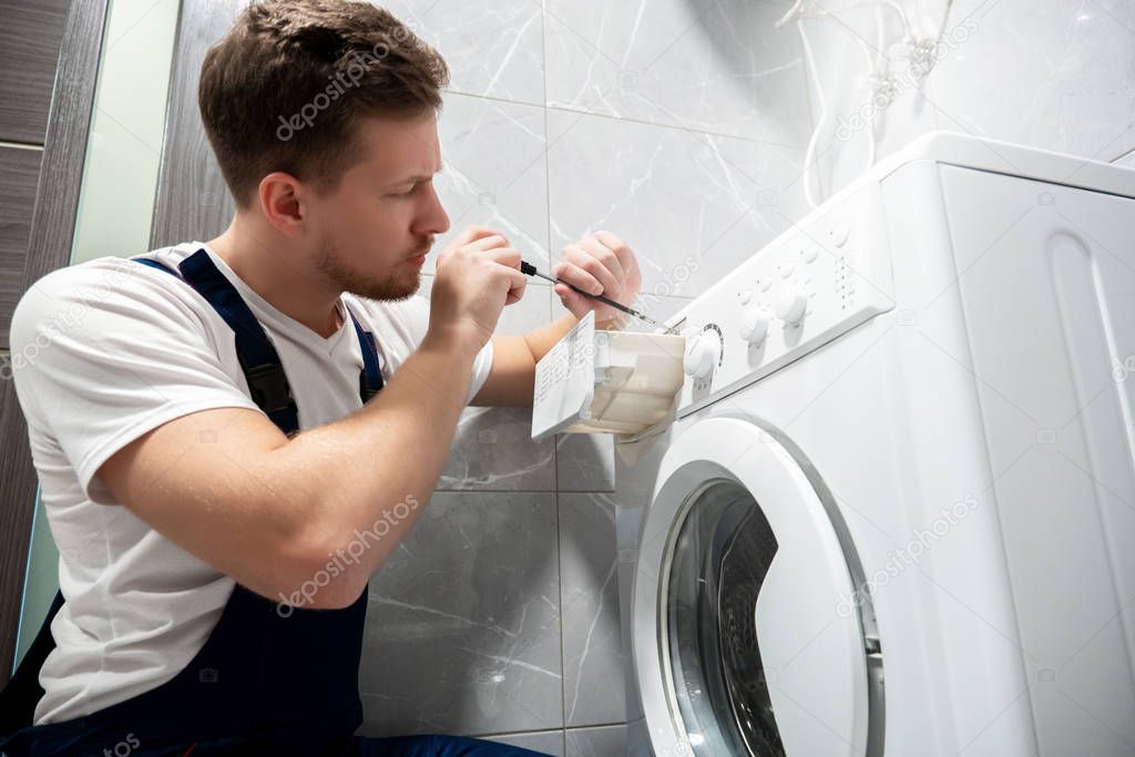 young handsome man worker in uniform repairing washing machine at home in the toilette looks concentrated professional repair service
