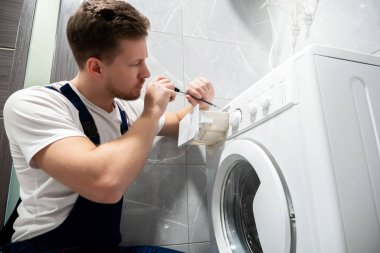Evde tuvalette çamaşır makinesini tamir eden üniformalı genç yakışıklı adam yoğun profesyonel tamir servisi gibi görünüyor.